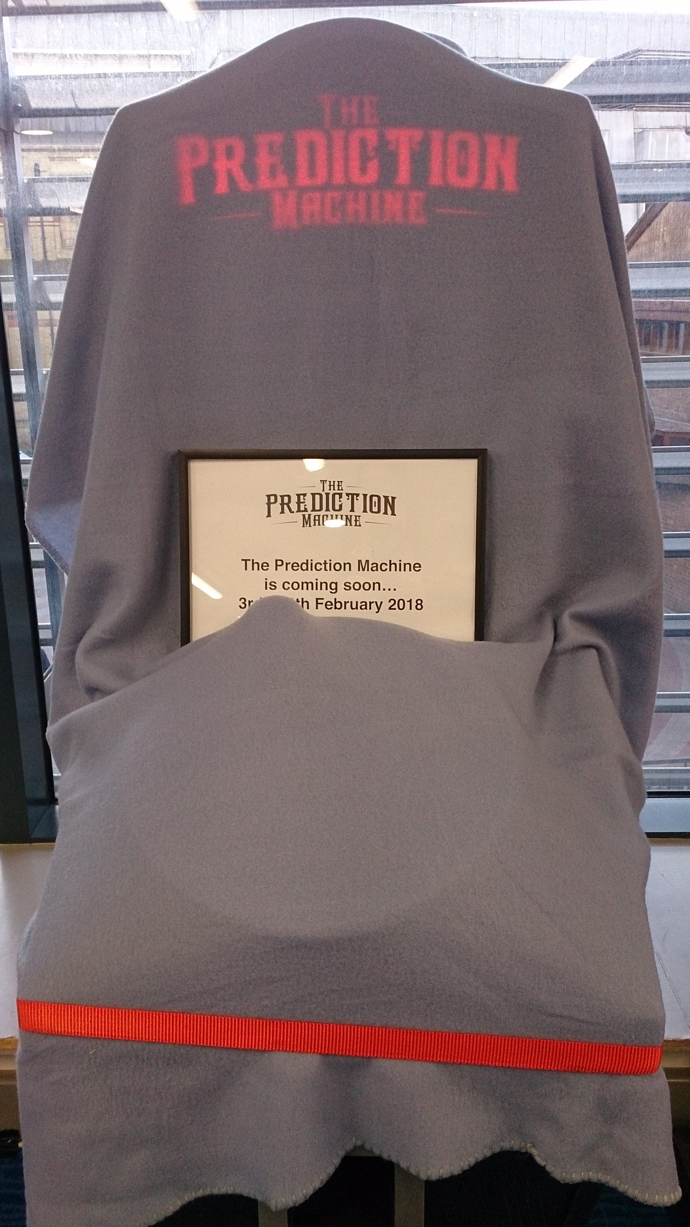 The Prediction Machine covered in a blanket with a sign saying it is coming soon before the exhibition started at Cambridge Central Library