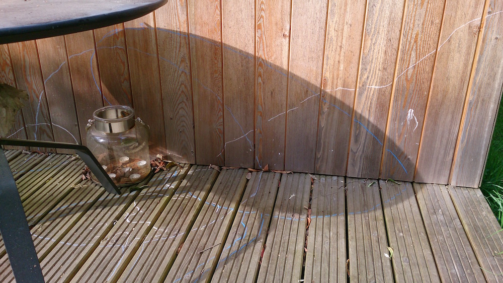 shadow of the round table and white lines drawn on the decking and wooden wall of the studio